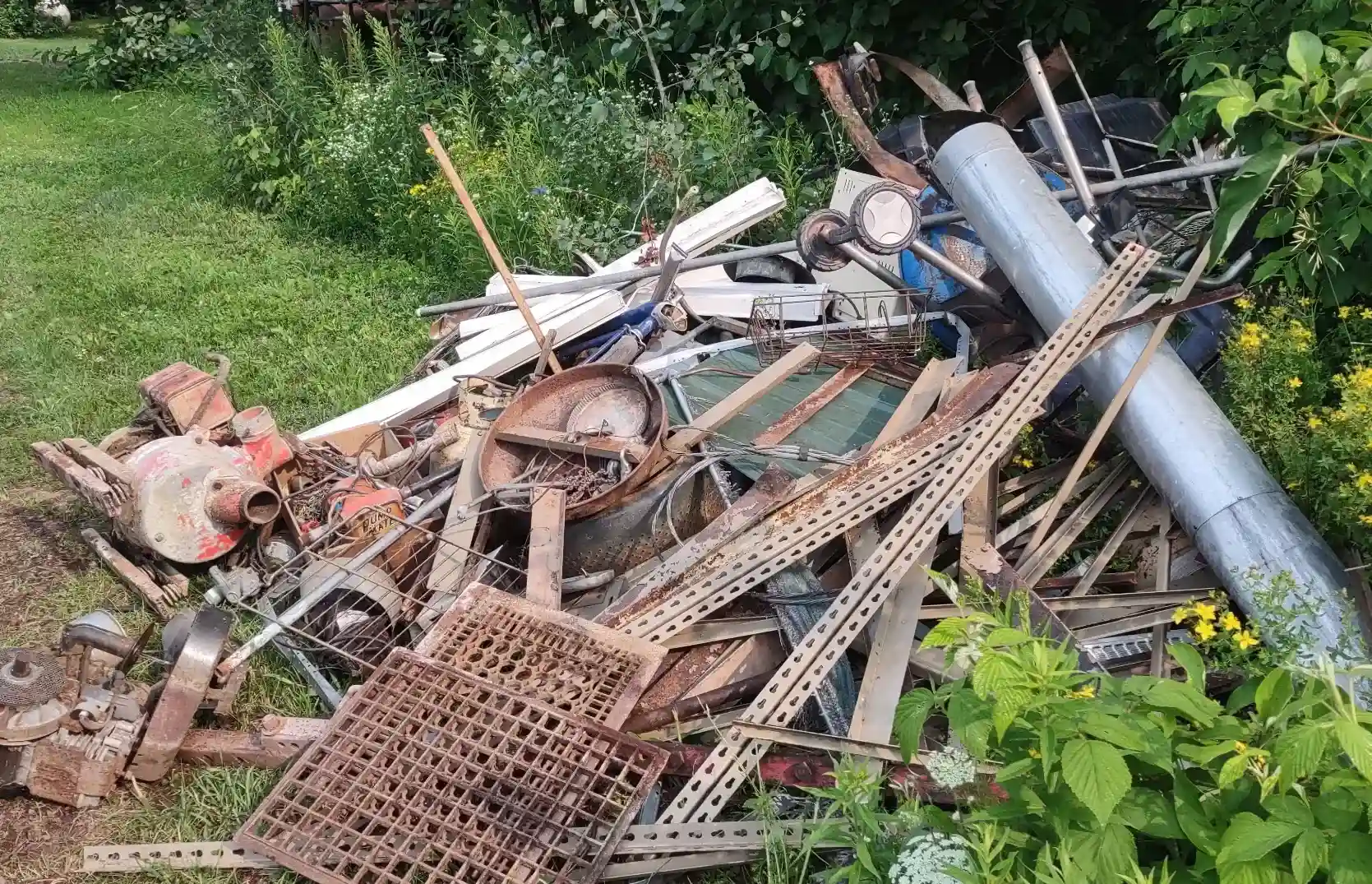 Overgrown pile of rusty metal scrap, pipes, and construction debris cluttering a grassy backyard before junk removal service